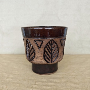 Peut inclure: Un pot de fleurs en céramique avec un bord et une base brun foncé brillants. La section centrale présente une finition texturée brun clair avec des motifs de feuilles et de triangles en relief. Le récipient a une large ouverture.