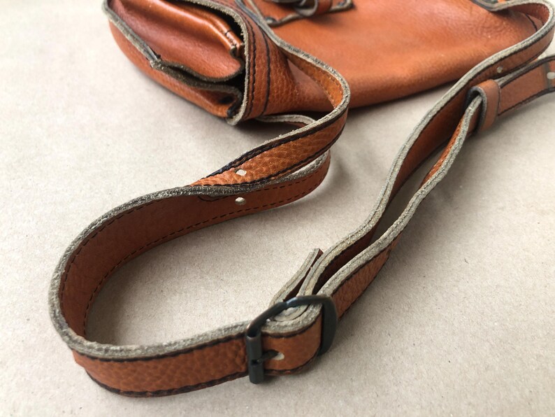 May include: A brown leather crossbody bag with a strap that has a buckle closure. The strap is adjustable and has a light brown trim.