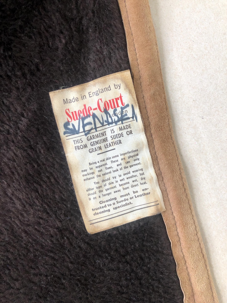 Puede incluir: Una etiqueta blanca con texto negro unida a una chaqueta de ante marr&oacute;n. La etiqueta dice "Hecho en Inglaterra por Suede-Court SVENDSEN. Esta prenda est&aacute; hecha de ante o cuero genuino. Al ser una piel real, algunas imperfecciones pueden ser evidentes. Estas son marcas f&iacute;sicas y no defectos, y solo realzan el aspecto natural de la prenda. Debes intentar evitar usar este tipo de piel con tiempo h&uacute;medo, pero si la prenda se moja, s&eacute;cala en una percha lejos del calor directo. La limpieza debe confiarse a un especialista en ante o cuero."