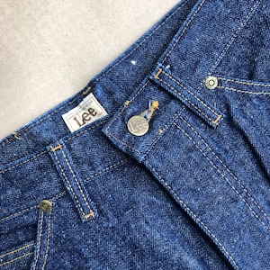 Könnte beinhalten: Nahaufnahme einer blauen Jeanshose mit einem braunen Leder-Lee-Etikett und einem Messingknopf. Die Jeans hat einen klassischen geraden Schnitt.
