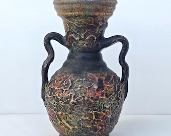 May include: A decorative vase with two curved handles, featuring a textured black surface with mottled patches of red, yellow, and green paint. The vessel has a wide, flared rim and a rounded body, showcasing a rugged, uneven finish throughout.