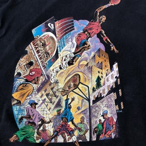 Może przedstawiać: Czarny T-shirt z kolorowym nadrukiem graficznym przedstawiającym koszykarzy w miejskim otoczeniu. Grafika przedstawia koszykarza w powietrzu, kosz do koszyk&oacute;wki i kilku innych graczy na boisku. Na grafice widoczny jest napis "Nike".