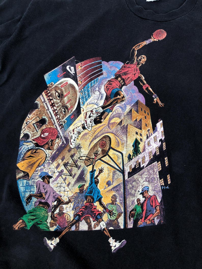 Może przedstawiać: Czarny T-shirt z kolorowym nadrukiem graficznym przedstawiającym koszykarzy w miejskim otoczeniu. Grafika przedstawia koszykarza w powietrzu, kosz do koszyk&oacute;wki i kilku innych graczy na boisku. Na grafice widoczny jest napis "Nike".