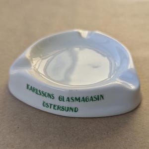 Puede incluir: Cenicero de cerámica blanca con forma triangular redondeada. El cenicero tiene una depresión central y tres apoyos curvos para cigarrillos. Texto verde en el lateral: "KARLSSONS GLASMAGASIN ÖSTERSUND".
