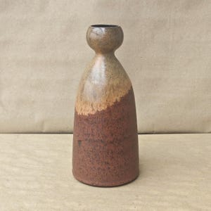 May include: A ceramic vase with a bulbous neck and a wide, cylindrical base. The upper portion features a smooth, light tan glaze, while the lower section is finished in a textured, dark reddish-brown glaze with a visible speckled appearance.