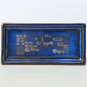 May include: A rectangular ceramic tray with a deep blue glaze and a dark brown rim. The center features a metallic-toned graphic illustration of trees, buildings, and the text GUSTAVSBERG. The tray has a raised border and a glossy finish.