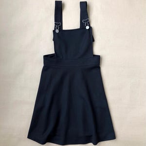May include: A solid black pinafore dress featuring a square neckline, wide shoulder straps, and silver-toned metal hardware clips that secure the straps to the front bodice. The skirt has a flared silhouette with a defined waistband.