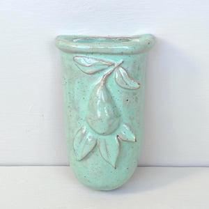 May include: A wall-mounted ceramic pocket vase in a light mint green glaze with a speckled finish. The front features a raised, molded relief of a single pear fruit with attached leaves and a stem, centered on the body of the vessel.