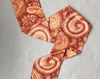May include: A long, thin scarf with a paisley pattern in shades of orange and brown. The scarf is folded in half and tied in a knot.