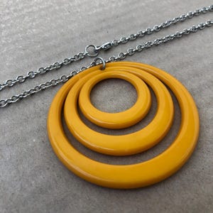 Op de afbeelding: Een gele ketting met een hanger met drie cirkels. De cirkels zijn concentrisch en gemaakt van een glad, glanzend materiaal.