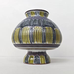 Pode incluir: Um vaso de cerâmica com corpo largo e arredondado e base de pedestal. A superfície apresenta um padrão repetitivo de formas verticais alongadas semelhantes a pétalas, alternando tons de cinza carvão escuro e verde oliva com acabamento brilhante.