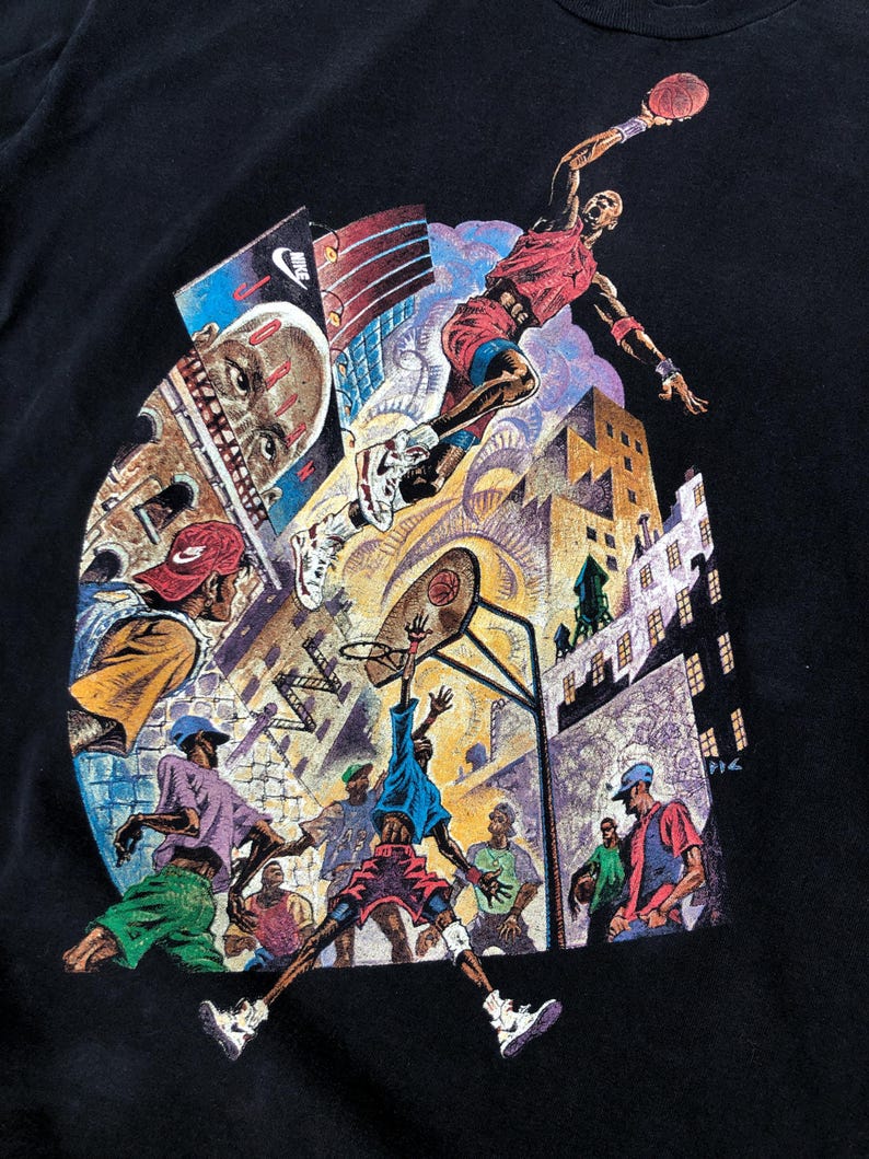 Może przedstawiać: Czarny T-shirt z kolorowym nadrukiem graficznym przedstawiającym koszykarzy w miejskim otoczeniu. Grafika przedstawia koszykarza w powietrzu, kosz do koszyk&oacute;wki i innych graczy na boisku. Grafika zawiera r&oacute;wnież budynki i inne elementy miejskie.