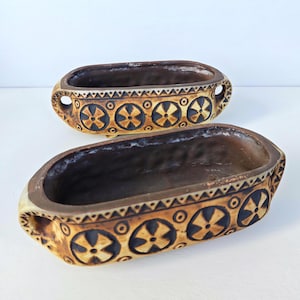 May include: Two oval ceramic planters with a dark brown interior and a tan exterior featuring a repeating pattern of dark brown circular motifs with four-pointed shapes and triangular borders, each with integrated side handles and small feet.