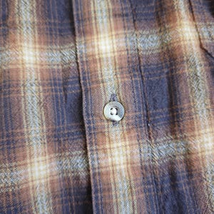 May include: Close-up of a button-down shirt with a plaid pattern in shades of brown, tan, and navy blue. The shirt features visible buttons and a textured fabric, suggesting a casual or workwear style.