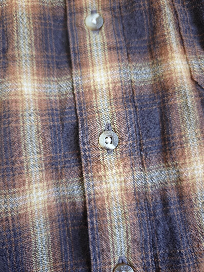 May include: Close-up of a button-down shirt with a plaid pattern in shades of brown, tan, and navy blue. The shirt features visible buttons and a textured fabric, suggesting a casual or workwear style.