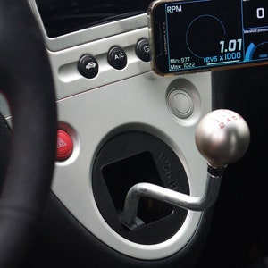 Könnte beinhalten: Nahaufnahme des Innenraums eines Autos, mit einem silbernen Schaltknauf mit roten Zahlen, einem schwarzen Lederlenkrad und einem Smartphone, das einen digitalen Drehzahlmesser mit einem Wert von 1,01 anzeigt.