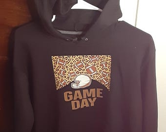 May include: Black hooded sweatshirt with a football-themed graphic. The graphic features a football helmet, footballs, and the words "GAME DAY" in brown. The background of the graphic has a leopard print design.