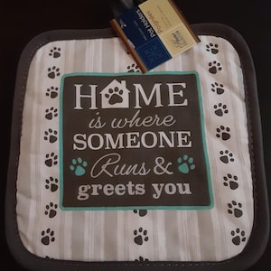 Puede incluir: Un agarrador de olla cuadrado con un borde gris y un fondo a rayas. El centro presenta el texto "HOME is where SOMEONE Runs & greets you" con huellas de patas. Huellas de patas adicionales están dispersas alrededor del borde.