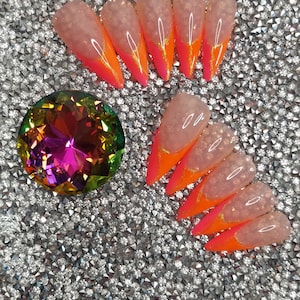 May include: A collection of artificial nails with a gradient design, transitioning from a clear base to a vibrant orange and pink tip. A large, multi-colored gemstone is also present, set against a sparkling, silver background.