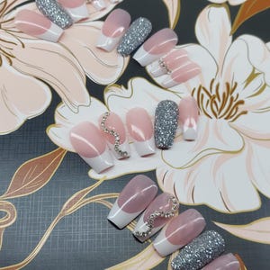 May include: A collection of artificial nails with a French manicure design. The nails feature a white tip, a pink base, and some are adorned with silver glitter or rhinestones. The nails are arranged on a floral patterned background.