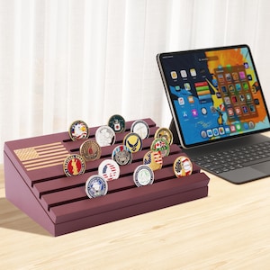 May include: A burgundy display stand with an American flag design, holding a collection of military challenge coins. The coins feature various emblems and designs. A tablet with a colorful screen is in the background.