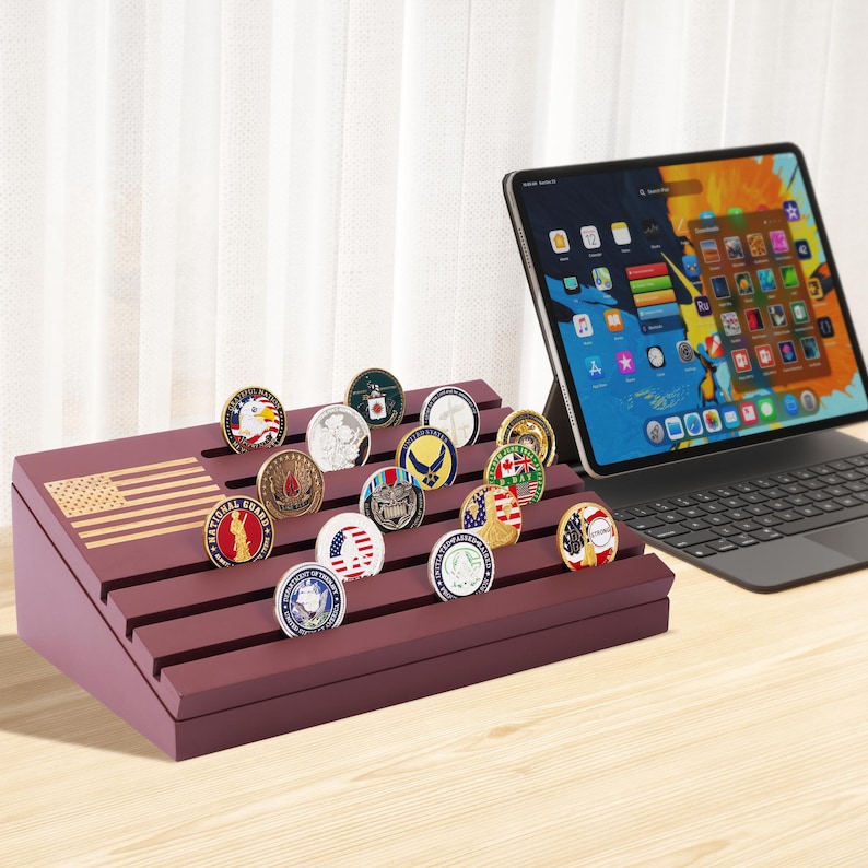 May include: A burgundy display stand with an American flag design, holding a collection of military challenge coins. The coins feature various emblems and designs. A tablet with a colorful screen is in the background.
