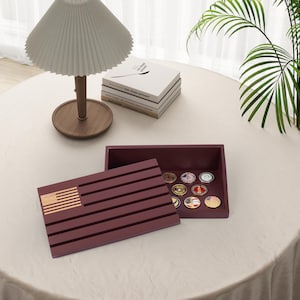 May include: A burgundy wooden box with an American flag design on the lid, open to reveal a collection of military challenge coins. The box is on a white tablecloth, with a lamp and books in the background.