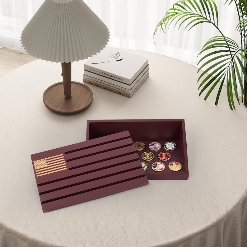 May include: A burgundy wooden box with an American flag design on the lid, open to reveal a collection of military challenge coins. The box is on a white tablecloth, with a lamp and books in the background.