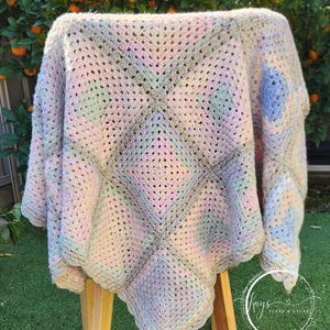May include: A pastel-coloured, handmade crochet blanket. The blanket has a granny square design in shades of pink, blue, green, and grey. It is draped over a wooden stand, with a green lawn and orange tree in the background.
