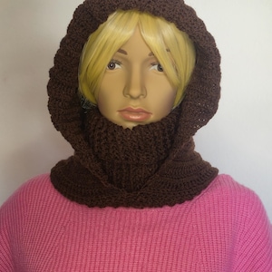 Könnte beinhalten: Ein braunes gehäkeltes Kapuzen- und Kragenset. Die Kapuze rahmt das Gesicht ein und ist mit einem Kragen verbunden, der den Hals bedeckt. Das Set wird auf einer Schaufensterpuppe mit einem rosa Pullover präsentiert.