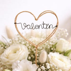 Könnte beinhalten: Ein Blumenstrauß aus weißen Rosen und anderen Blüten, gekrönt von einem herzförmigen Aufsatz mit dem Schriftzug "Valentin" in Schreibschrift. Der Aufsatz besteht aus Draht und Bindfaden.