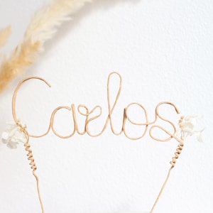 Könnte beinhalten: Handgefertigter Tortenaufsatz mit dem Namen "Carlos" in Schreibschrift aus beigefarbenem Draht. Kleine weiße Blumen und dekorative Drahtakzente verzieren die Basis. Der Hintergrund ist eine schlichte weiße Oberfläche.