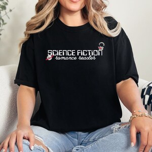 May include: A woman wearing a black t-shirt with the text "SCIENCE FICTION romance reader" and a small rocket ship graphic. She is sitting on a white couch with a black and white checkered throw blanket.