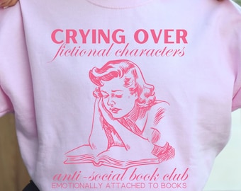 Könnte beinhalten: Hellrosafarbener Pullover mit dem Text "CRYING OVER fictional characters anti-social book club EMOTIONALLY ATTACHED TO BOOKS" und einer rosa Illustration einer Frau, die ein Buch liest.