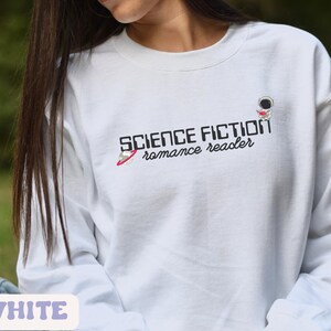 Puede incluir: Sudadera blanca de manga larga con el texto "SCIENCE FICTION romance reader" y una pequeña ilustración de un astronauta leyendo un libro.