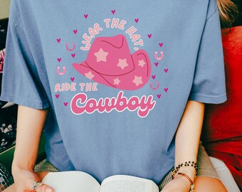Peut inclure: Un t-shirt bleu jean avec un graphique de chapeau de cowboy rose et le texte "Wear the Hat, Ride the Cowboy".