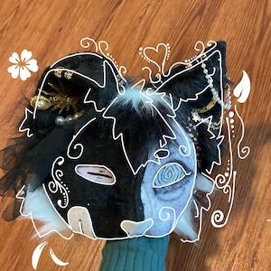 May include: A black and white cat-themed mask with intricate detailing. The mask features a split design, with one side black and the other a light blue. Embellishments include pearls, gold accents, and white fur.