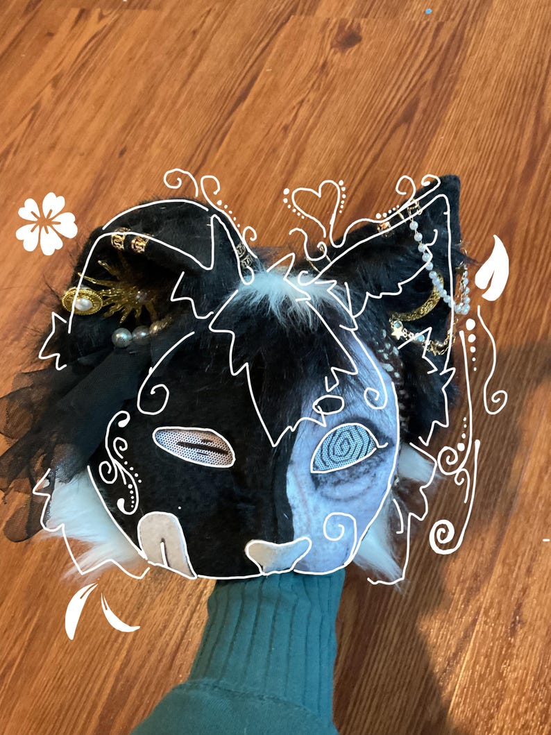 May include: A black and white cat-themed mask with intricate detailing. The mask features a split design, with one side black and the other a light blue. Embellishments include pearls, gold accents, and white fur.