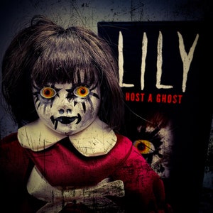 May include: A creepy doll with dark hair, yellow eyes, and a painted face, wearing a red dress. The doll is positioned in front of a black book cover that reads "LILY HOST A GHOST". The image has a distressed, vintage aesthetic.