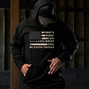 May include: Black hooded sweatshirt with a graphic design resembling the American flag. The design includes a wrench and the text "THAT'S WHAT I DO, I FIX STUFF, AND I KNOW THINGS" in white lettering. The sweatshirt has a front pocket.