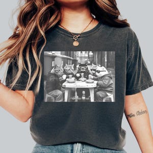 May include: Black and white graphic tee featuring a group of cats in hoodies and sunglasses, gathered around a small table, eating. The image is printed on a dark gray t-shirt.