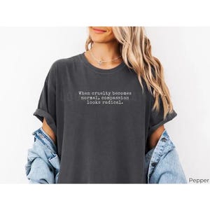 May include: Dark gray t-shirt with rolled-up sleeves. The shirt features the text: "When cruelty becomes normal, compassion looks radical." A denim jacket is worn over the shirt.