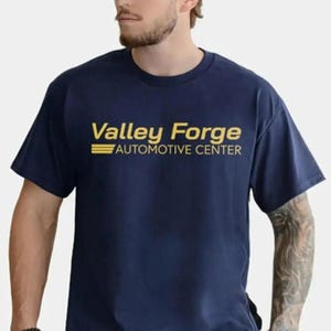 May include: Navy blue t-shirt with the text "Valley Forge AUTOMOTIVE CENTER" in gold. The shirt has short sleeves and a crew neck. The text is centered on the chest.