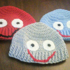 May include: Three handmade crochet hats in red, blue, and gray. Each hat features a smiling face with white eyes, black pupils, and a red smile. The hats are designed for children.