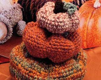 May include: A stack of three crocheted pumpkins in shades of orange, brown, and green. The pumpkins have a textured appearance and are arranged in a tiered fashion, with the largest at the base and the smallest at the top. The top pumpkin has a brown stem and green leaves.