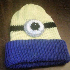 May include: A knitted beanie hat in yellow and blue, featuring a large eye design. The hat has a blue ribbed brim, a yellow crown, and a black stripe across the middle. The eye is white, gray, and black.