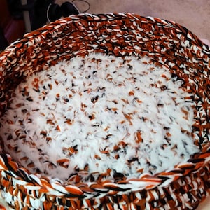May include: A round, crocheted pet bed with a white, fluffy lining. The bed is made with orange, black, and white yarn.