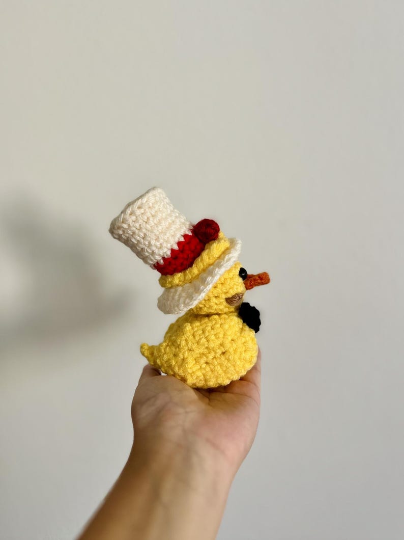 May include: A yellow crocheted duck wearing a white and red crocheted top hat with a black bow tie.