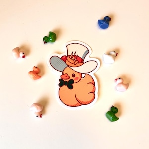 May include: An orange duck sticker wearing a top hat and bow tie, surrounded by small, colorful duck figurines. The sticker has a cartoon style, with a white outline and a pop art style background. The figurines are in various colors.