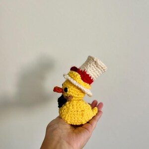 May include: A yellow crocheted duck wearing a white and red top hat. The duck is sitting in the palm of a hand.